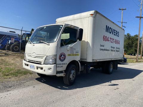 A 2014 Hino 195 box truck with a white cargo area and a company logo on the side parked on a road