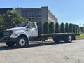 A white 2004 Ford F-750 flatbed truck with a yellow light on the roof parked on a paved surface with a row of trees in the bed