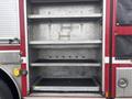 A red 2006 E-One Typhoon fire truck has an open storage compartment with three metal shelves and hooks for equipment