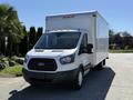 A 2015 Ford Transit with a white cargo box and black front grille parked with its headlights on