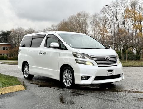 A white 2009 Toyota Vellfire is parked at an angle showcasing its sleek design and alloy wheels
