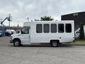 A white 2014 Ford E-350 van with large side windows is parked, featuring a simple design and a high roof suitable for passenger transport