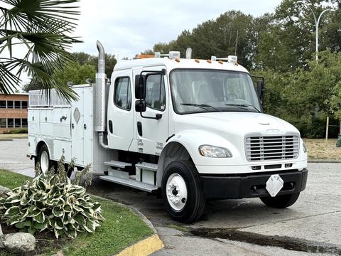 A white 2005 Freightliner M2 106 truck with a utility body and side compartments parked on a roadway
