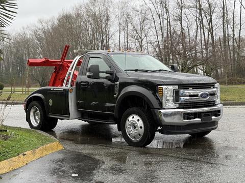 A black 2018 Ford F-450 Super Duty tow truck with a red tow mechanism in the bed is parked on a wet surface