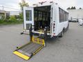 A 2021 Ford Transit bus with a ramp lowered at the rear for accessibility, featuring multiple seats and a white exterior