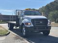 A white 2004 Ford F-750 flatbed truck with a mesh back and orange warning light on the roof positioned slightly angled toward the viewer