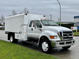 A 2013 Ford F-650 truck with a white body and a large cargo box attached to the bed is parked on the street